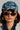 Wearing the DENIM SNAPBACK with a textured finish and adjustable buckle strap, along with dark oval sunglasses, large hoop earrings, and a white jacket, the person with long dark hair gazes directly at the camera against a plain background.