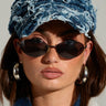 Wearing the DENIM SNAPBACK with a textured finish and adjustable buckle strap, along with dark oval sunglasses, large hoop earrings, and a white jacket, the person with long dark hair gazes directly at the camera against a plain background.