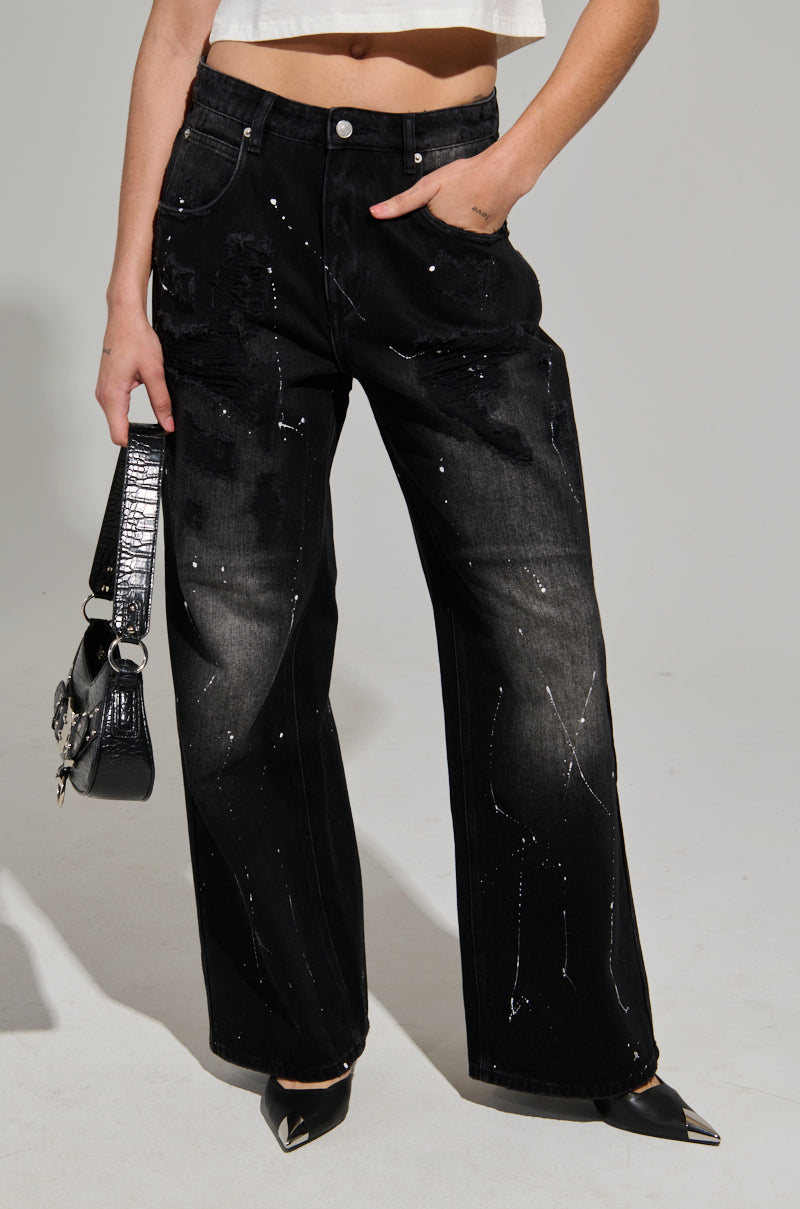 A person wearing the BACK IN BLACK DISTRESSED DENIM JEANS, featuring a high-waisted, wide-leg silhouette with white paint splatters, paired with a cropped white top. They're holding a black handbag and sporting pointed black shoes against a minimalistic neutral background.