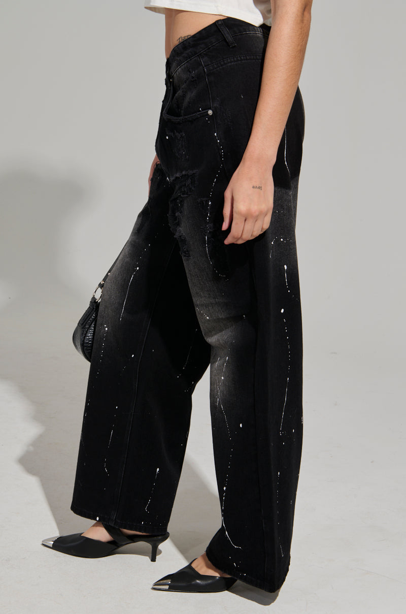 The individual is wearing the BACK IN BLACK DISTRESSED DENIM JEANS featuring a wide-leg cut and white splatter pattern, complemented by a white crop top and black slingback heels. They are holding a black handbag while posed against a plain gray background.
