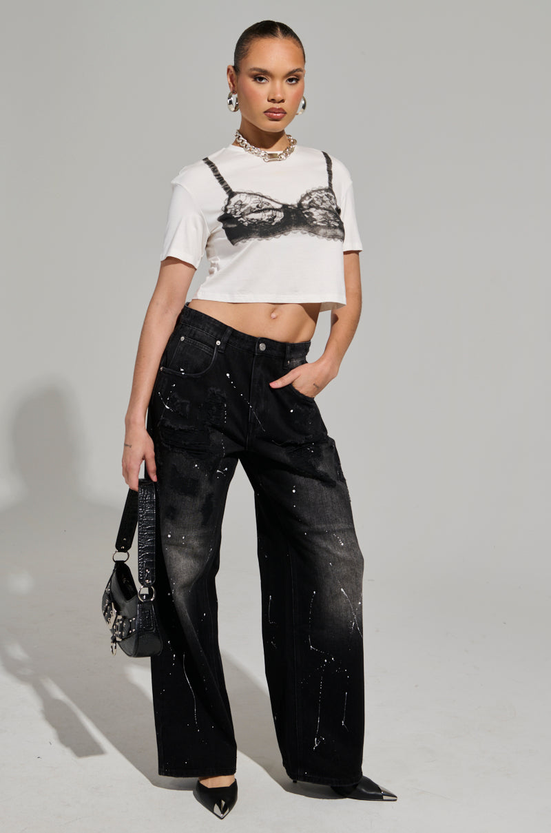 A person poses confidently in a fashion-forward outfit: a cropped white t-shirt with a printed black lace design, BACK IN BLACK DISTRESSED DENIM JEANS with wide legs and a subtle splatter pattern emphasizing the silhouette, pointed black shoes, and a small black handbag.
