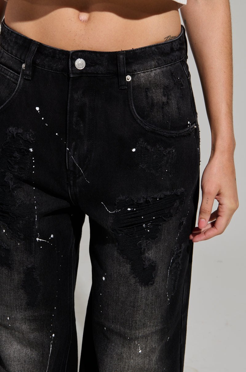 A person wearing the BACK IN BLACK DISTRESSED DENIM JEANS, featuring white paint splatters and stylish rips. These dark wash denim jeans have a button closure, perfectly capturing the trend. Only the lower torso and upper legs are visible against a plain background.