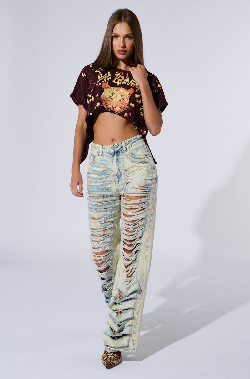 A woman poses in a casual outfit featuring a cropped Def Leppard t-shirt and the OVER IT DISTRESSED HIGH RISE JEANS. She complements her look with leopard print shoes and stands against a plain white background.