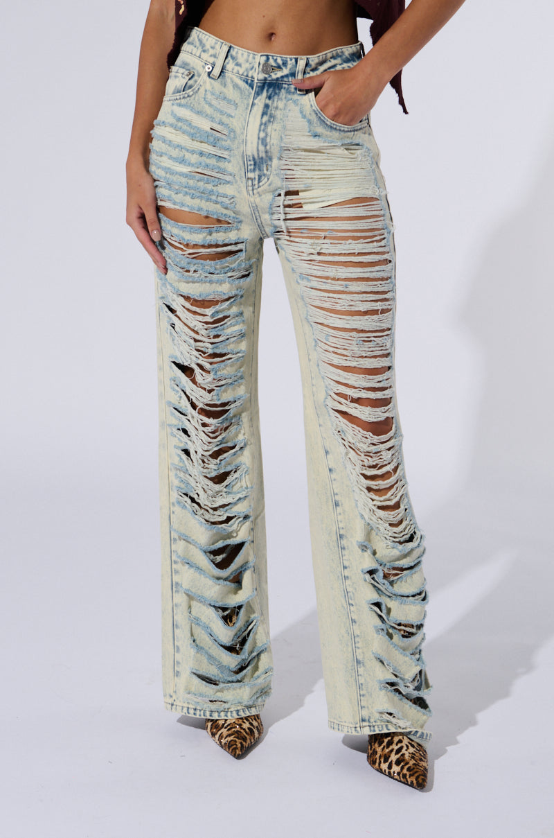 A person stands with their right hand in their pocket, wearing OVER IT DISTRESSED HIGH RISE JEANS made from cotton light wash denim. The extensive rips run from the upper thighs to the ankles, paired perfectly with leopard print shoes. The jeans feature a straight leg silhouette against a plain white background.