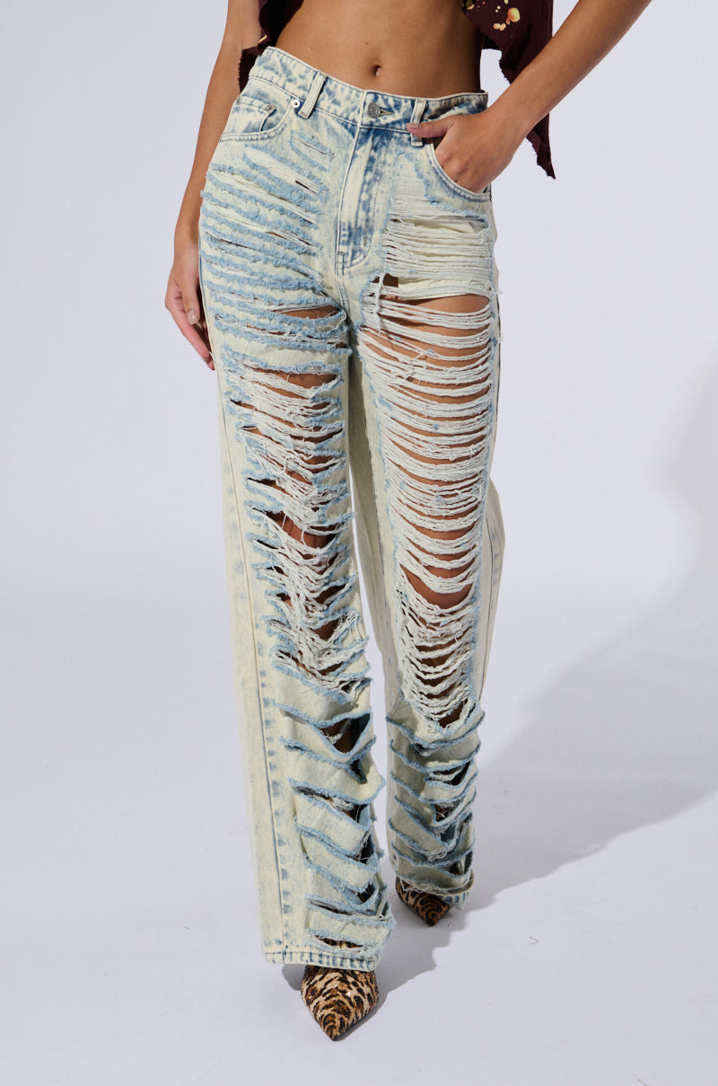 A person is shown from the waist down wearing a pair of Over It Distressed High Rise Jeans made from cotton light wash denim. The heavily ripped jeans with a straight-leg silhouette feature extensive horizontal tears revealing the legs underneath. The person wears pointed leopard print shoes and has one hand in a pocket.