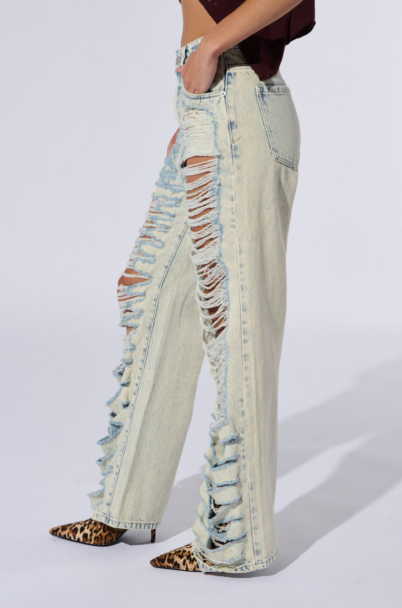 A person is wearing the OVER IT DISTRESSED HIGH RISE JEANS, which feature a light-wash, heavily distressed design with large rips and fraying from the mid-thigh to the ankle. They are also sporting pointed-toe leopard print shoes. The background is plain white.