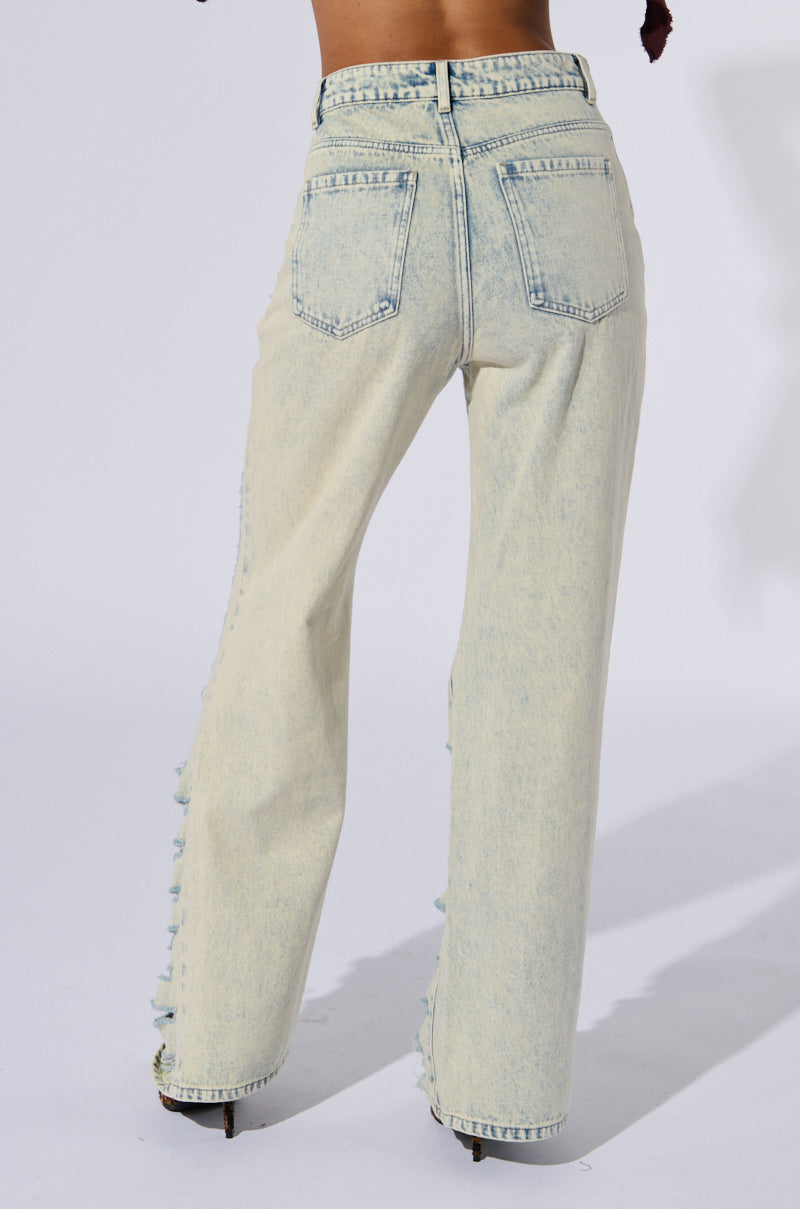 A person wearing OVER IT DISTRESSED HIGH RISE JEANS in cotton light wash denim with a straight leg silhouette and frayed details stands facing away from the camera. The jeans have two back pockets and are paired with a black top. The background is plain white.