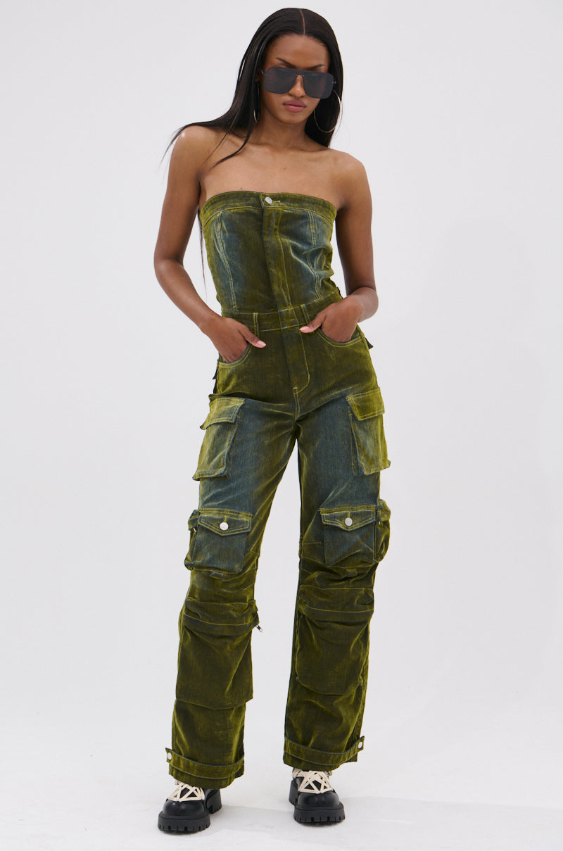 A woman channels tomboy chic in dark sunglasses and the MIRA DISTRESSED VELVET DENIM JUMPSUIT—strapless, olive green, with cargo pockets—paired with chunky black-and-beige shoes, hands-in-pockets against a plain white background.