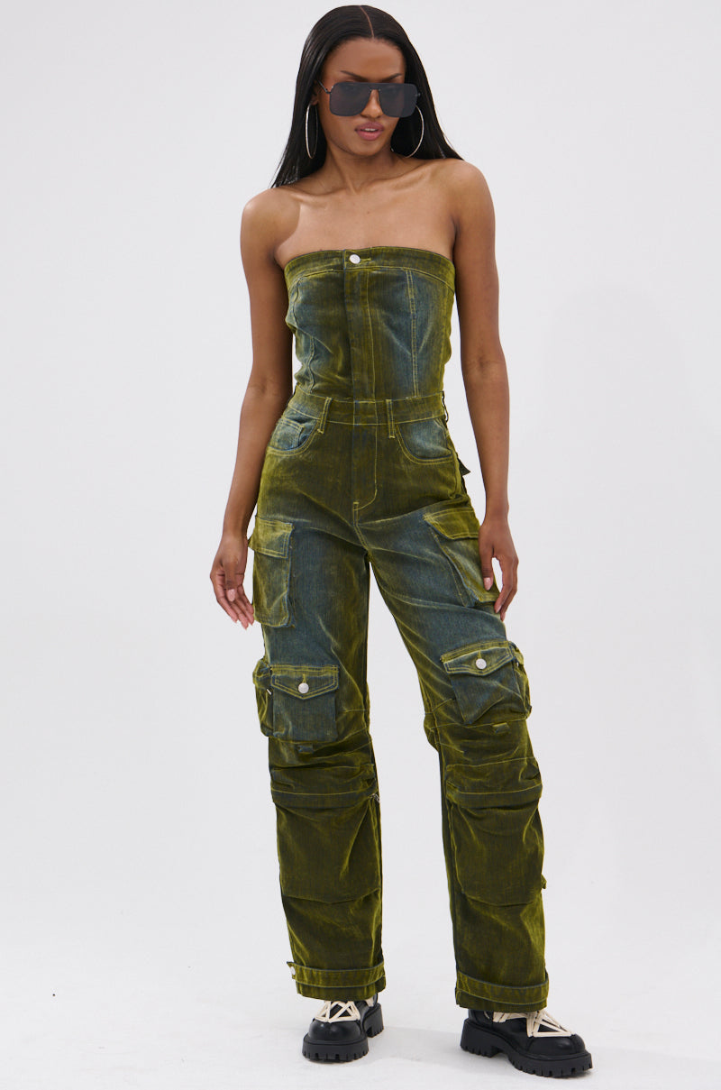 A woman stands against a white background in the MIRA DISTRESSED VELVET DENIM JUMPSUIT, styled with black chunky shoes, large hoop earrings, and oversized sunglasses for a tomboy chic look.