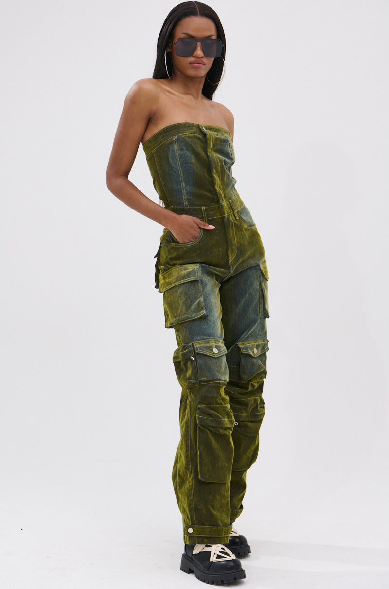 A woman in oversized sunglasses and hoop earrings poses in the MIRA DISTRESSED VELVET DENIM JUMPSUIT—a strapless, greenish-yellow piece with cargo pockets—styled with black chunky shoes for a tomboy-chic look against a white background.