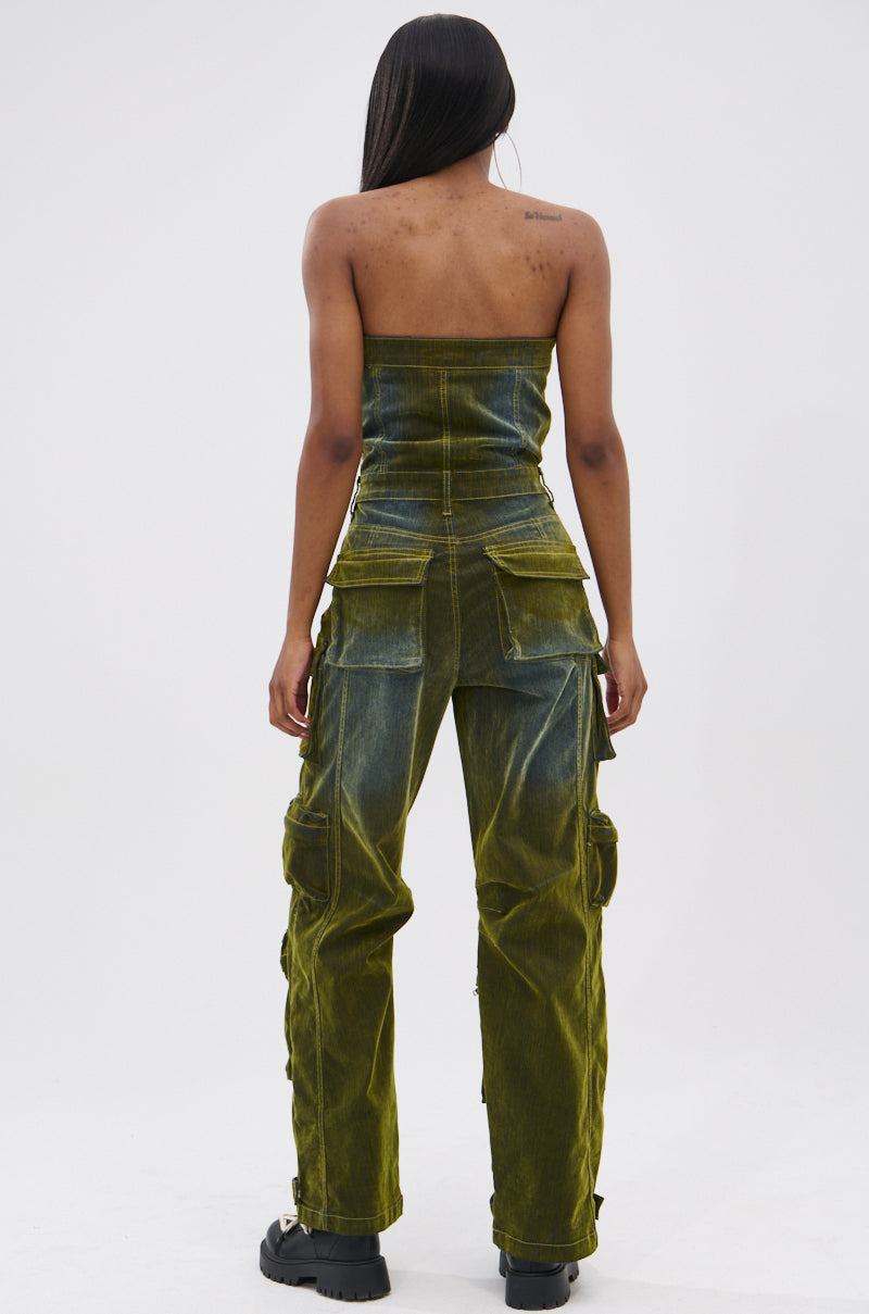 A person with long hair faces away, wearing the MIRA DISTRESSED VELVET DENIM JUMPSUIT—strapless, green, with large pockets—paired with chunky black shoes for a tomboy chic look against a plain white background.