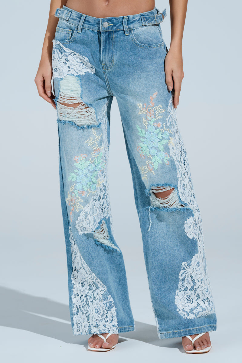 A model wears the PICKING PETALS FLORAL AND LACE DETAIL DENIM PANTS with distressed rips, white lace, a high waist, and a loose fit, styled with open-toe heels against a light blue background.