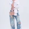 A woman poses against a white background wearing a white off-the-shoulder blouse, PICKING PETALS FLORAL AND LACE DETAIL DENIM PANTS, white open-toe heels, and holding a pink patterned handbag.