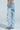 A person in profile wears PICKING PETALS FLORAL AND LACE DETAIL DENIM PANTS—light blue, mid-rise, distressed jeans with large white lace patches—paired with open-toe sandals against a light blue background for an airy look.