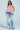 A woman faces forward in a strapless pink peplum top and PICKING PETALS FLORAL AND LACE DETAIL DENIM PANTS with a mid-rise fit and distressed details. She has wavy brown hair and wears white heeled sandals.