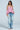 A woman with wavy, shoulder-length brown hair poses against a plain light background wearing PICKING PETALS FLORAL AND LACE DETAIL DENIM PANTS, styled with a strapless ruffled pink top.
