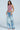 A woman poses in PICKING PETALS FLORAL AND LACE DETAIL DENIM PANTS—mid-rise, light blue, distressed with white lace accents—paired with a sleeveless pink halter top. She stands against a plain background, arms relaxed and softly smiling.
