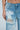A close-up shows a person wearing PICKING PETALS FLORAL AND LACE DETAIL DENIM PANTS—mid-rise blue jeans with a large rip and white floral lace applique by the pocket and hip. Their hand rests by their side against a light blue background.