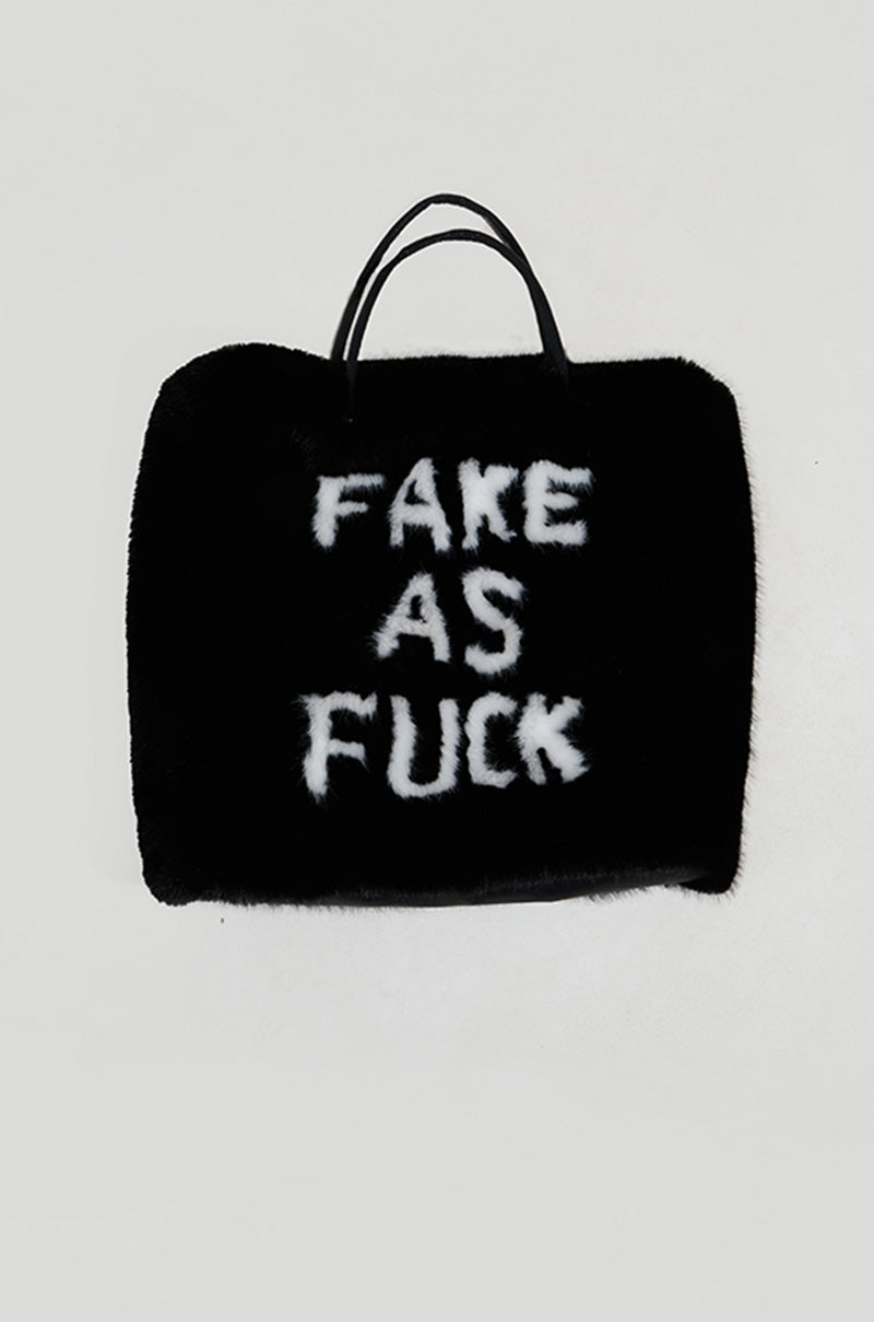 Introducing the IM FAKE ULTRA OVERSIZED FUR TOTE: A black oversized fur tote boldly emblazoned with the phrase "FAKE AS FUCK" in white letters. The bag showcases two sleek faux leather handles and presents a roomy satin-lined interior, set against a plain light gray backdrop to hold all your essentials.