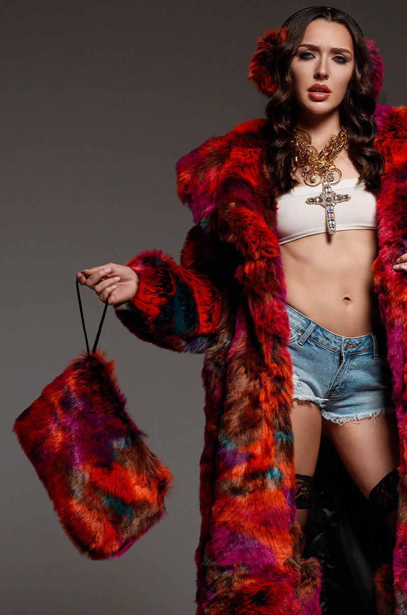 A woman showcases a vibrant multicolor faux fur coat and matching earmuffs, holding the chic MAXIMUS FUR WRISTLET in her left hand. She pairs it with a white crop top, denim shorts, and a bold necklace against a neutral gray background.