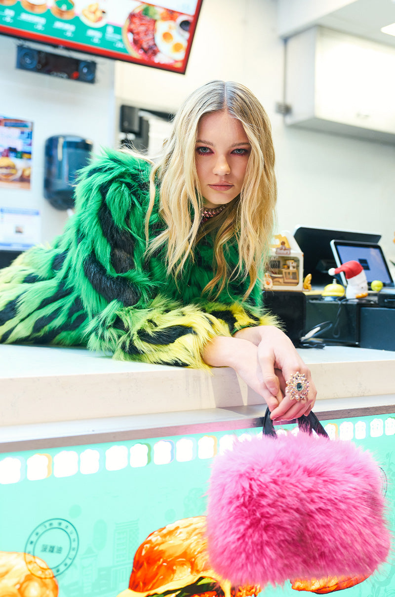 A person with long blonde hair leans on a counter, wearing a vibrant green and black furry jacket. They hold the SABRINA FAUX FUR BAG, which is embellished with faux leather handles. The background displays restaurant menus and a cash register, enhancing the lively atmosphere.