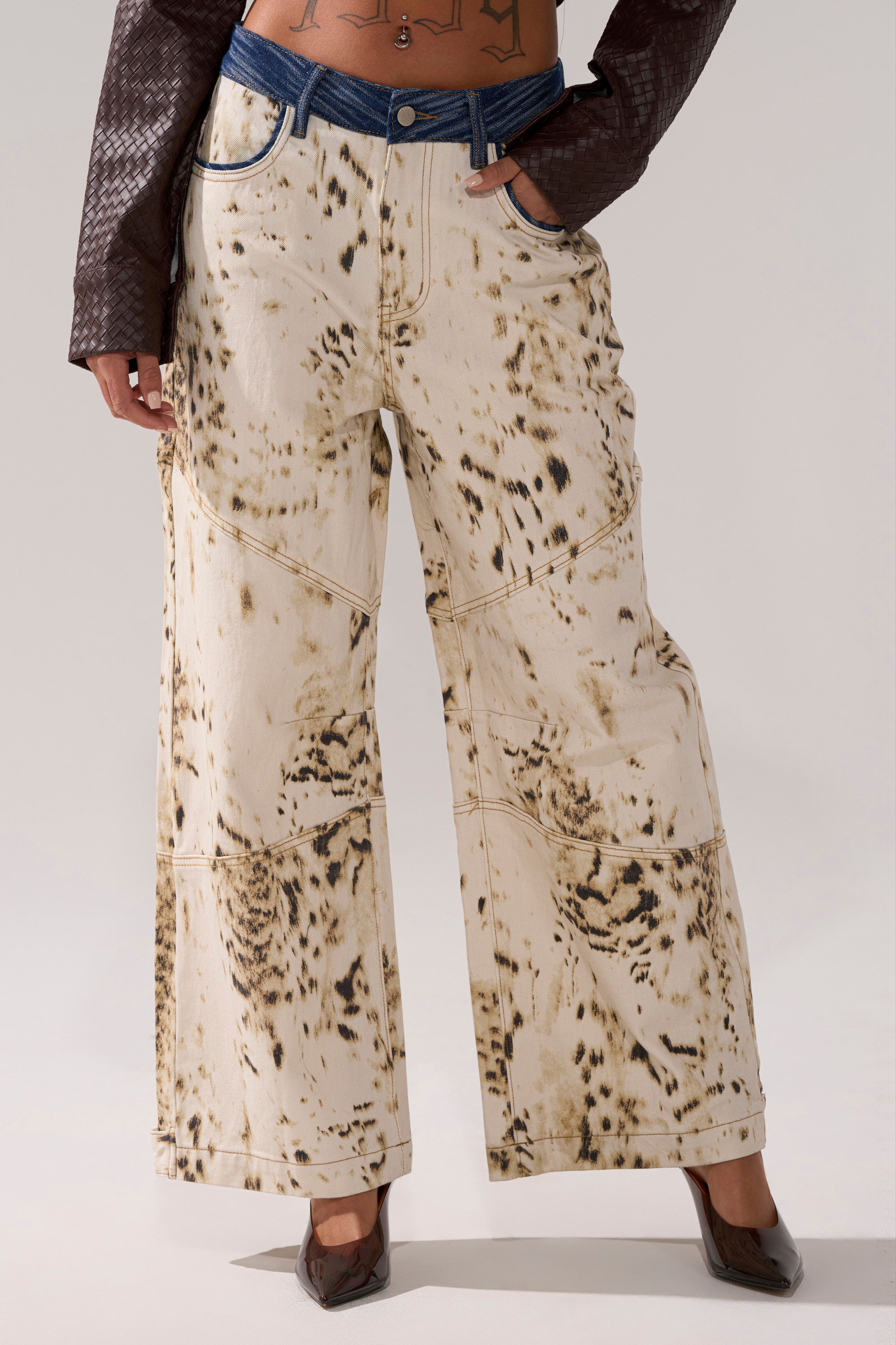 A person wears the BAMBI GIRLY WIDE LEG DENIM, featuring a beige and brown bleached pattern with a blue waistband, styled with pointed brown heels and one hand in their pocket. 0