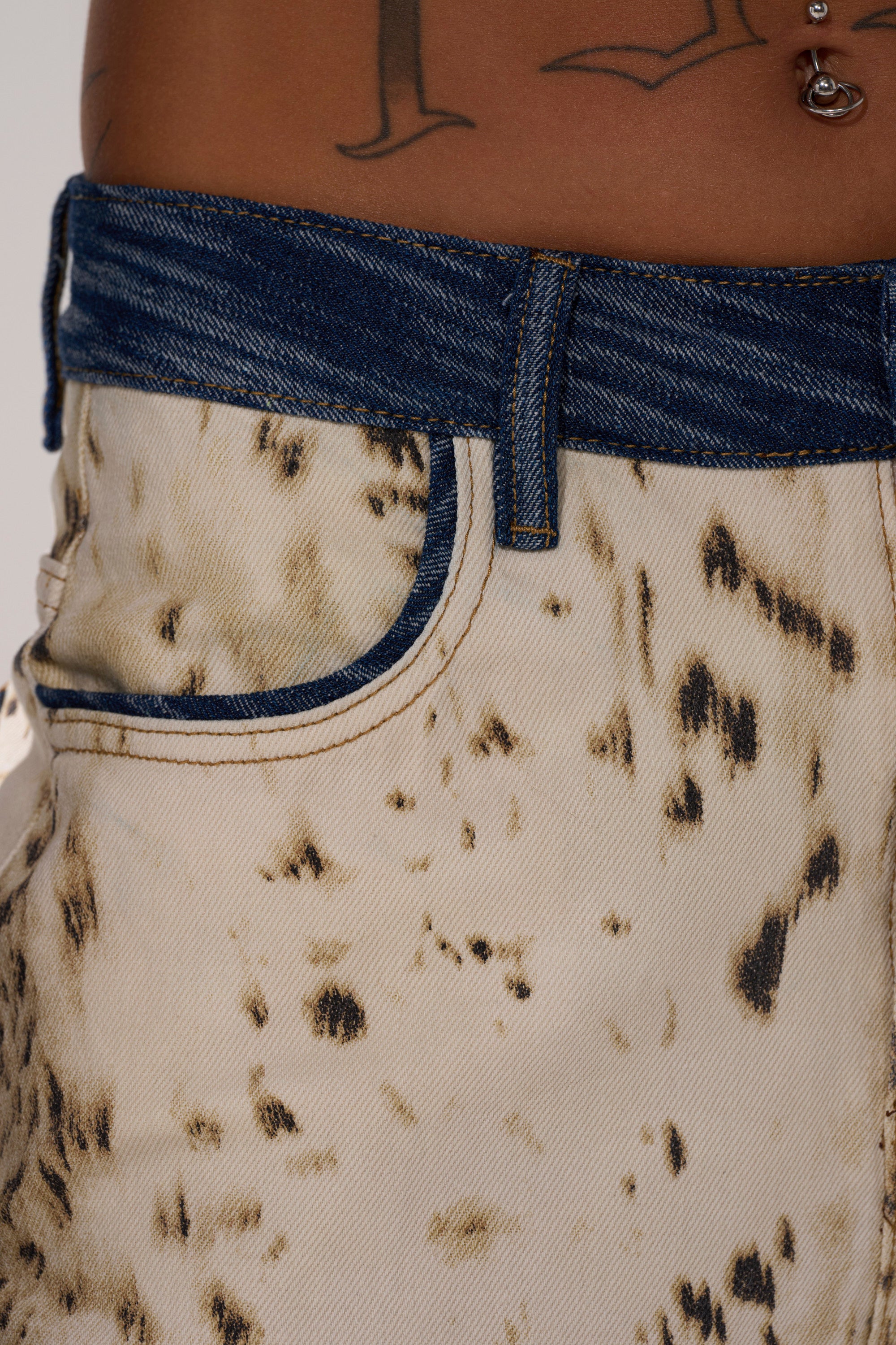 Close-up of a person wearing BAMBI GIRLY WIDE LEG DENIM, featuring a bleach-splattered pattern, blue waistband and trim, with a hint of tattoo and navel piercing visible above the jeans. 8