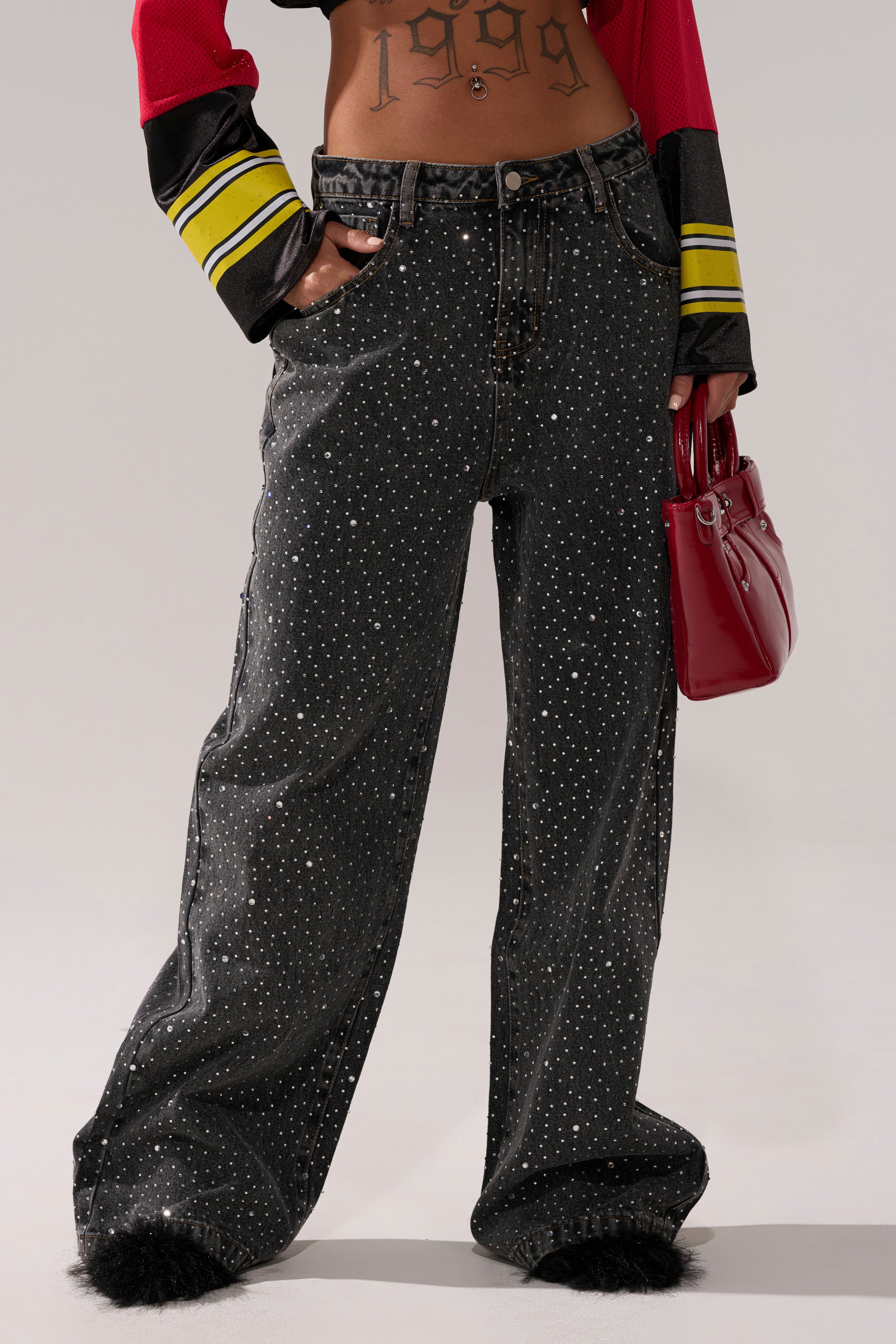A person wears RHINESTONE QUEEN EMBELLISHED DENIM—wide-leg, dark wash jeans with white polka dots and fur-trimmed hems—paired with a crop top, "1999" tattoo visible, holding a small red handbag. Their head is not in view.