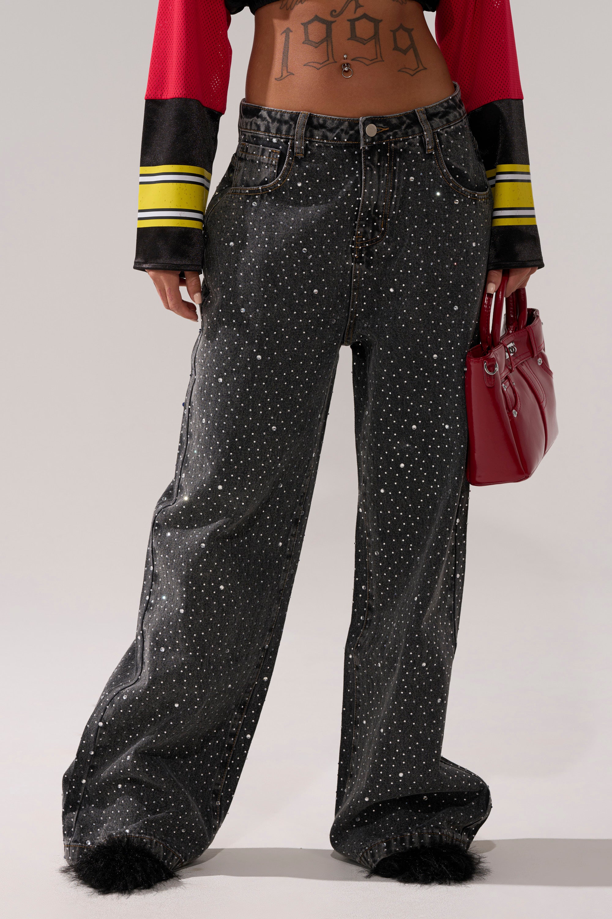 A person wearing RHINESTONE QUEEN EMBELLISHED DENIM stands with a red handbag, showing a "1999" midriff tattoo and sporting a red top with yellow and black striped sleeves.