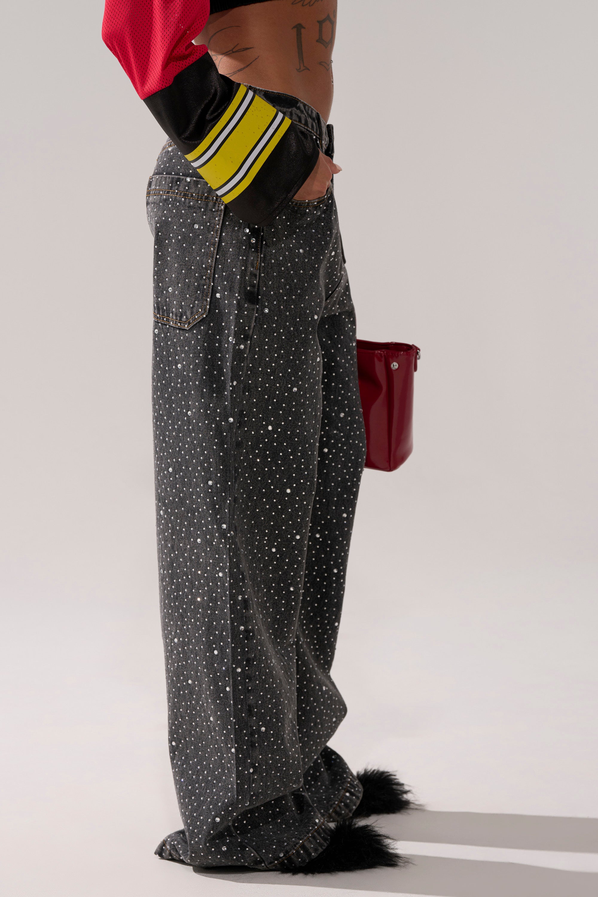 A person wears RHINESTONE QUEEN EMBELLISHED DENIM—wide-leg, dark-wash jeans featuring white dot patterns and black fur cuffs—paired with a red and black jacket and a shiny red bag, standing against a plain background.