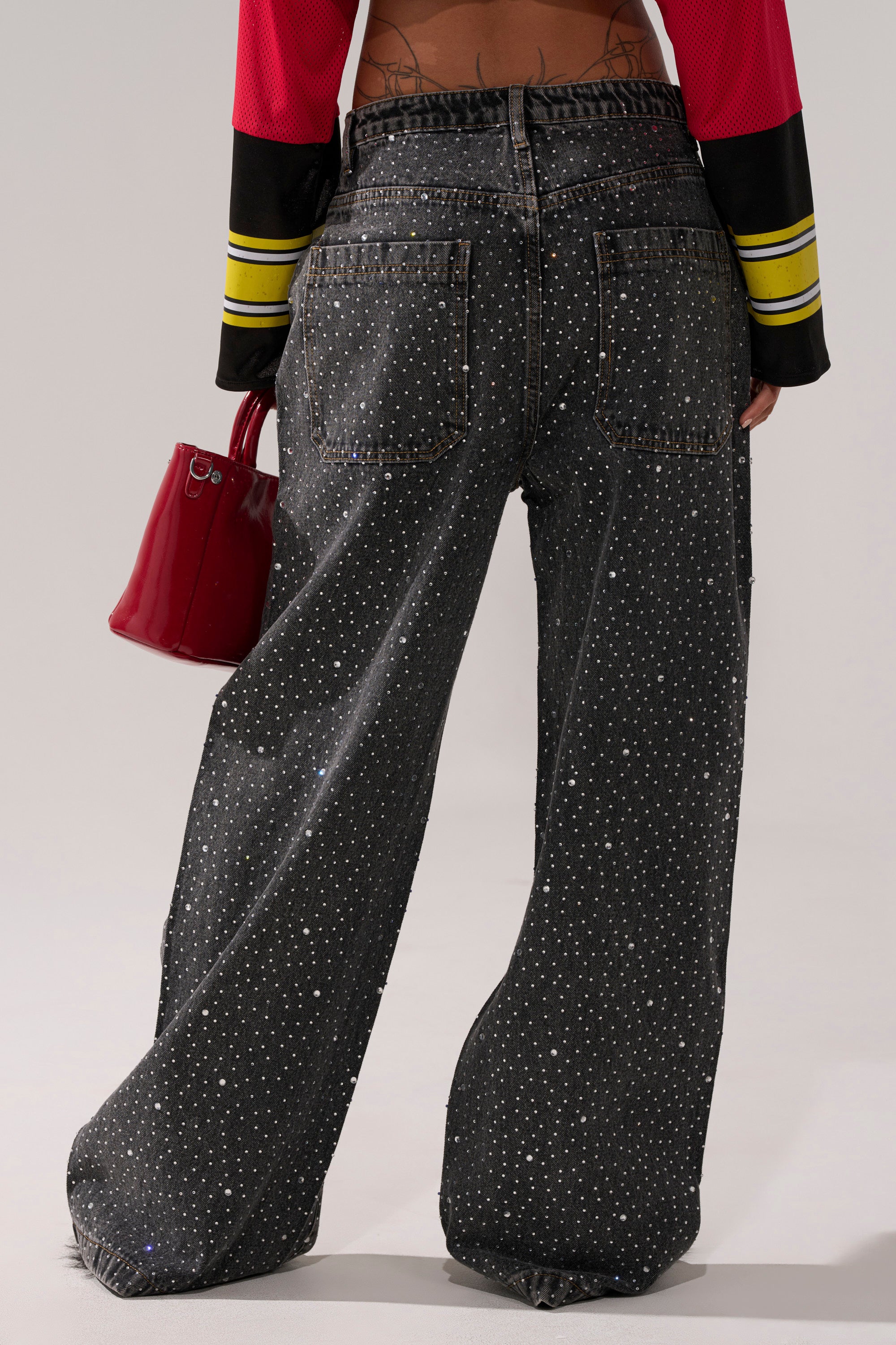 A person is shown from the back wearing RHINESTONE QUEEN EMBELLISHED DENIM wide-leg black jeans, a red top with black and yellow sleeves, and carrying a shiny red handbag.