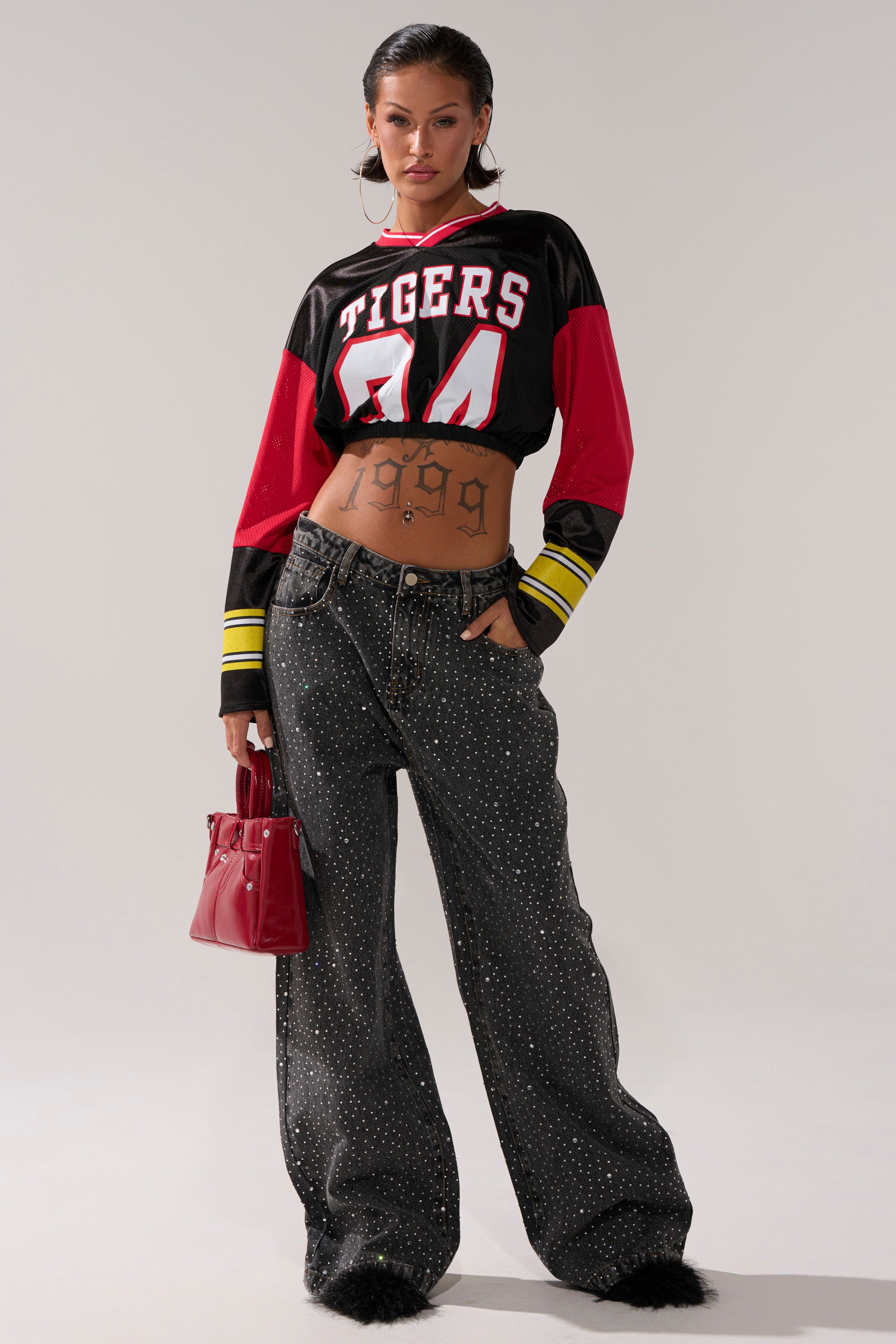 A woman wears a cropped "Tigers 00" jersey, RHINESTONE QUEEN EMBELLISHED DENIM, black shoes, hoop earrings, and holds a red handbag. A "1999" tattoo is visible on her stomach.