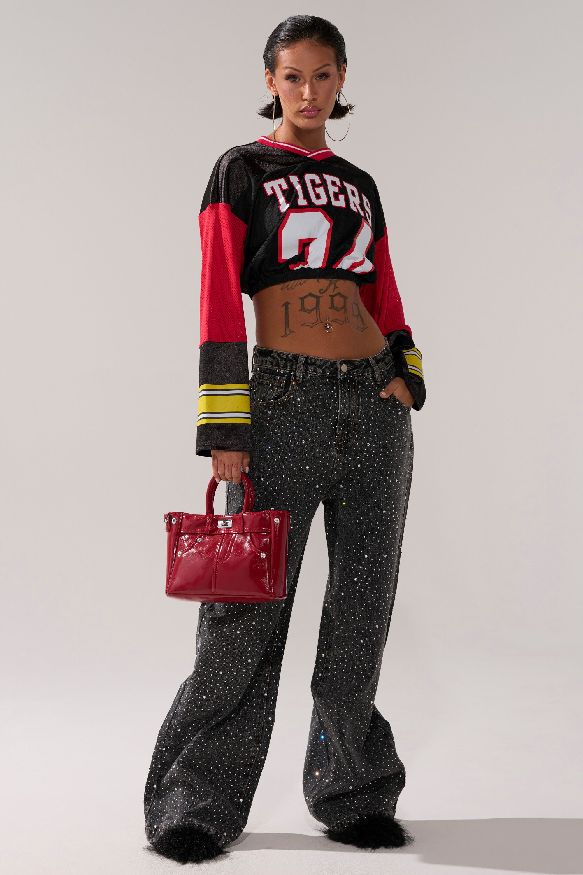 A woman stands confidently in a cropped "Tigers 04" sports jersey, wearing RHINESTONE QUEEN EMBELLISHED DENIM jeans, black furry shoes, large hoop earrings, and holding a shiny red handbag. Her midriff tattoo reads "1999".