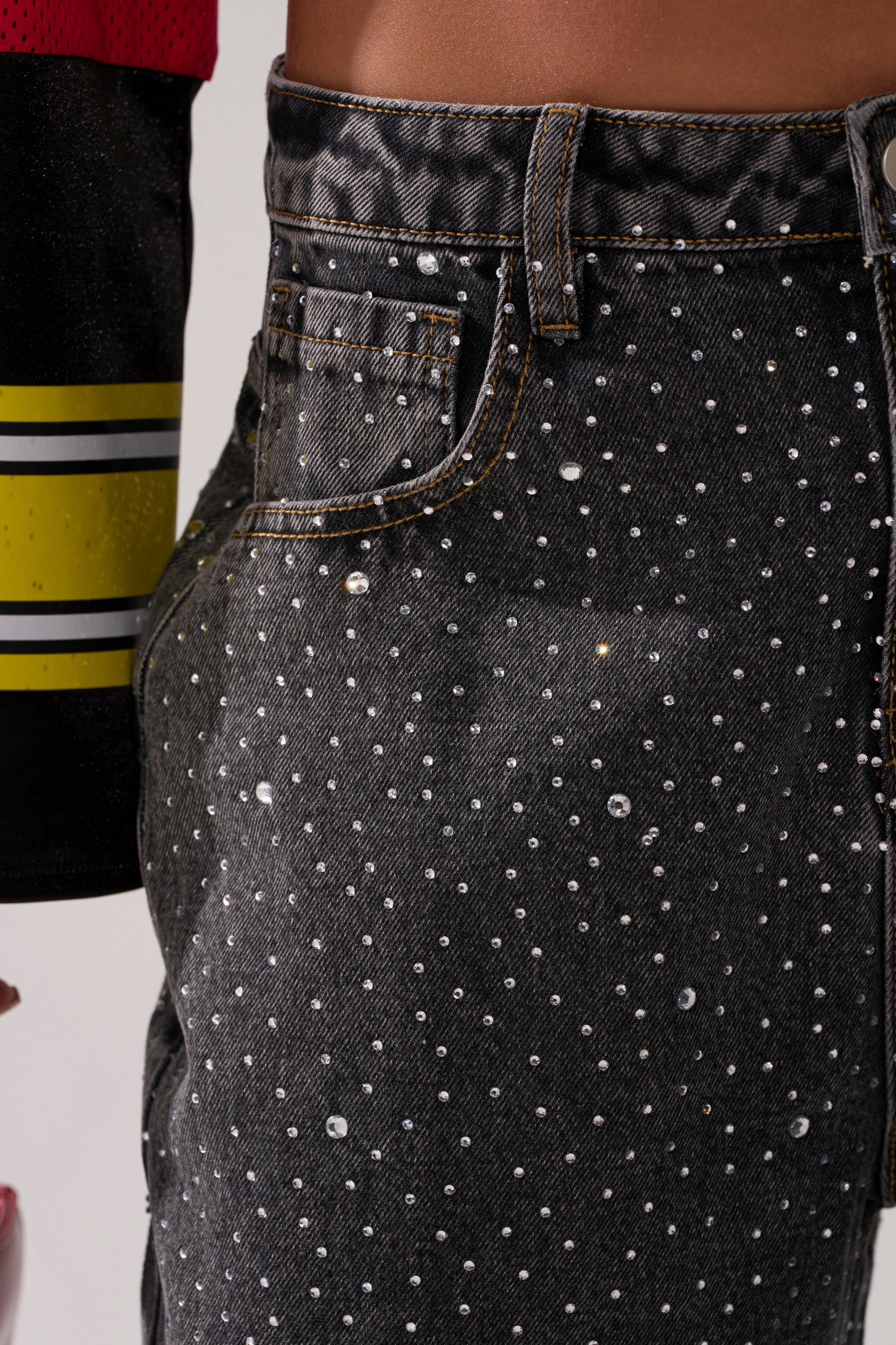 Close-up of a person's waist in the RHINESTONE QUEEN EMBELLISHED DENIM—black, dark-wash jeans adorned with small rhinestones. A black, red, and yellow striped sleeve appears at the left edge of the image.