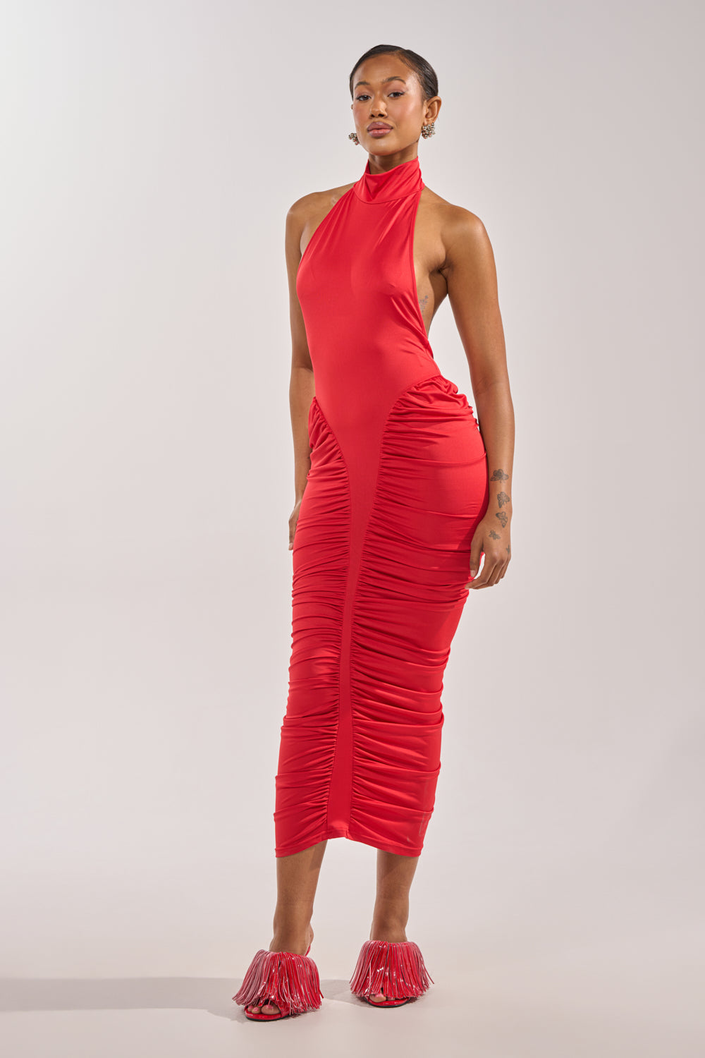 A woman exudes confidence in the HIPS DON'T LIE RUCHED MAXI DRESS IN RED, a sleeveless, high-neck style that echoes sporty varsity charm as she stands against a plain light background. 0