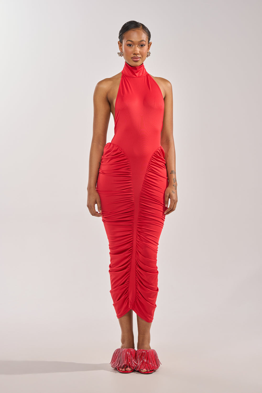 A person faces forward in the HIPS DON'T LIE RUCHED MAXI DRESS IN RED, a sleeveless, high-neck, fitted dress with ruching, styled with varsity-inspired red shoes featuring fringe accents against a plain light background. 1
