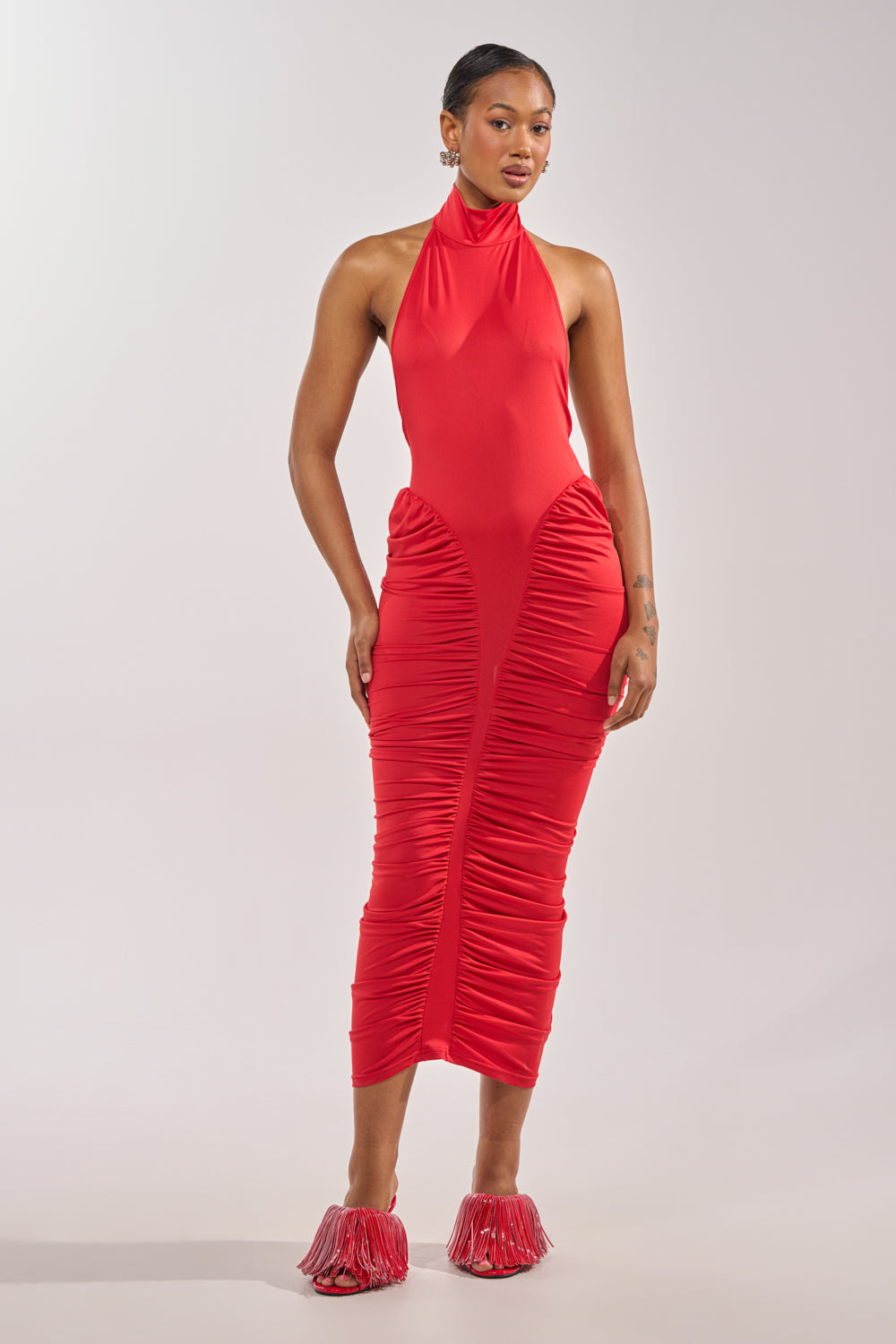 A woman wears the HIPS DON'T LIE RUCHED MAXI DRESS IN RED—sleeveless, halter-neck, form-fitting with ruched detail—paired with red fringed heels, standing against a plain light background for a striking look distinct from classic varsity styles. 2