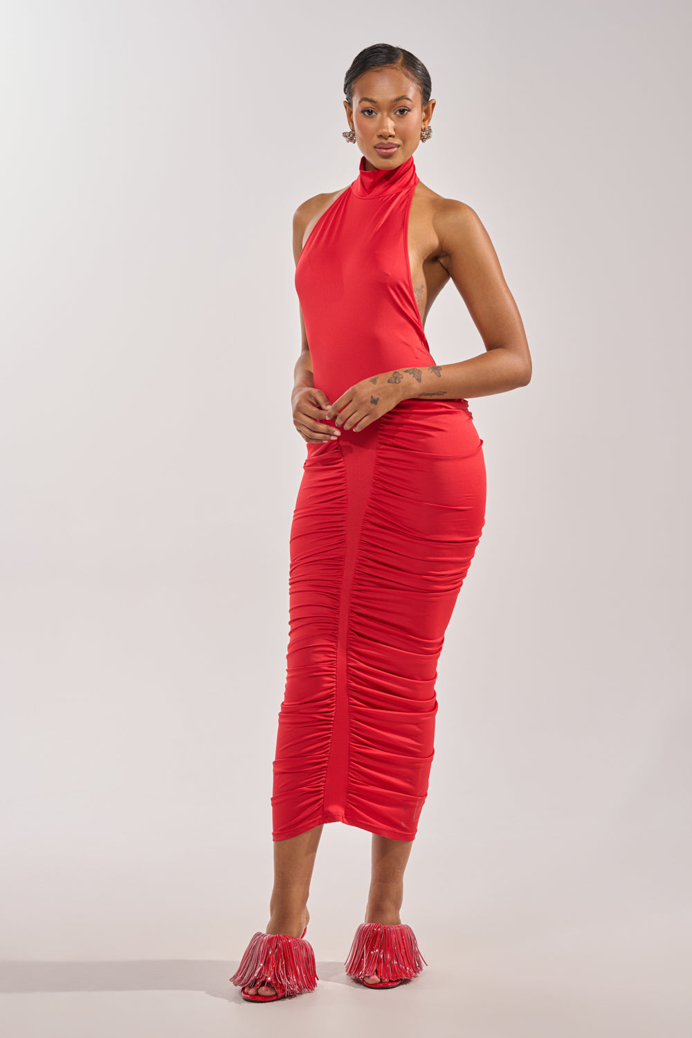 A woman stands confidently against a plain background, wearing the HIPS DON'T LIE RUCHED MAXI DRESS IN RED, a fitted sleeveless dress with front ruching, paired with red fringed heeled shoes, channeling bold varsity-inspired style. 3
