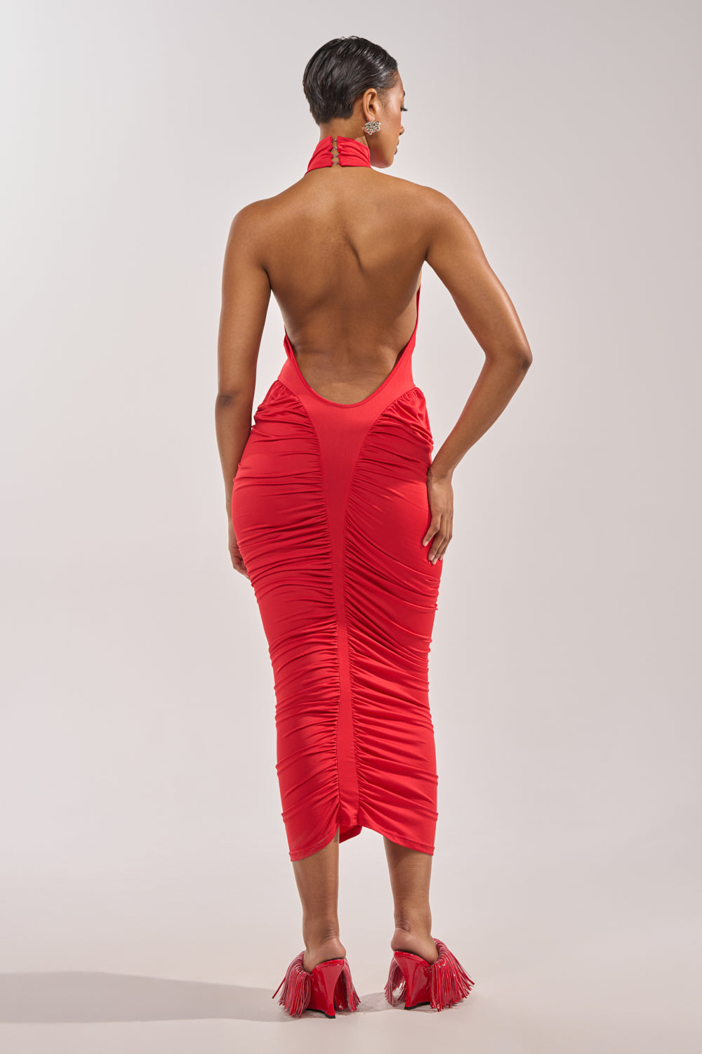 A woman stands with her back turned, wearing the HIPS DON'T LIE RUCHED MAXI DRESS IN RED and red heeled sandals. Her sleek short hair pops against a plain gray backdrop, blending boldness with sporty varsity shirt-inspired style. 5