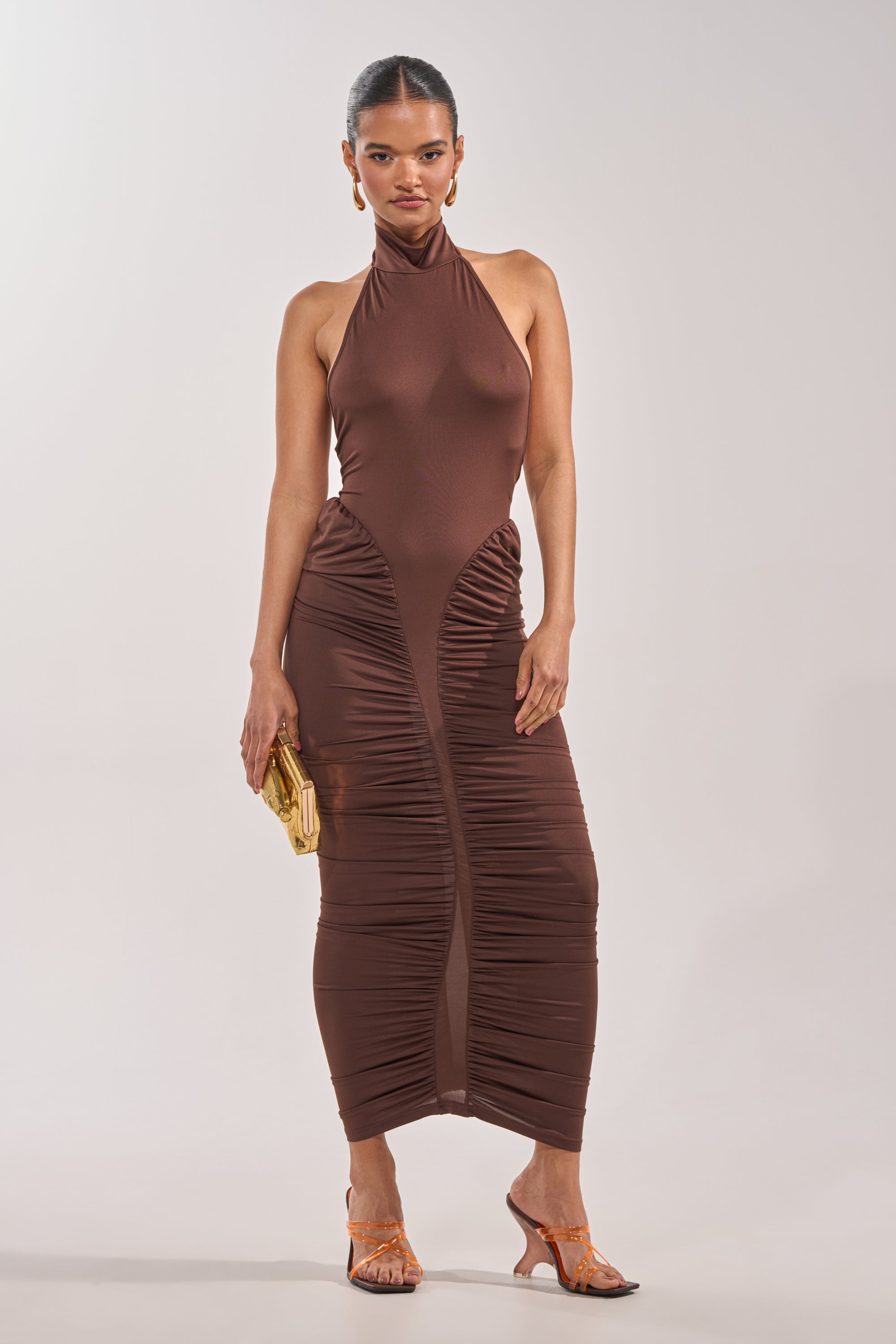 A woman models the HIPS DON'T LIE RUCHED MAXI DRESS in chocolate brown, featuring a sleeveless halter design and ruched details, styled with a gold clutch, orange heeled sandals, and gold hoop earrings. Ideal as a cocktail party dress. 1