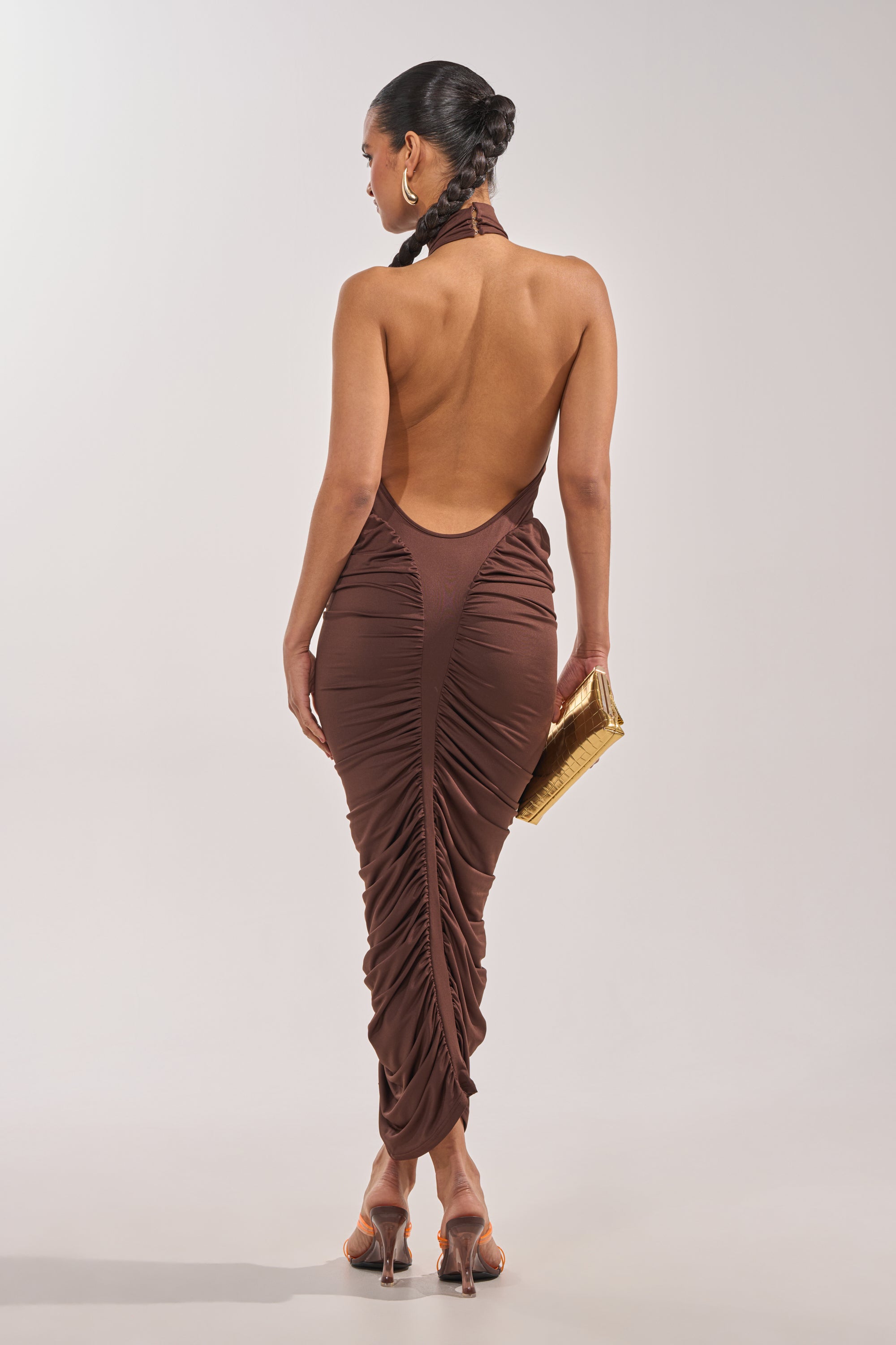 A woman poses with her back to the camera, wearing the HIPS DON'T LIE RUCHED MAXI DRESS in chocolate brown, heeled sandals, and a gold clutch, her hair styled in a sleek bun. 4