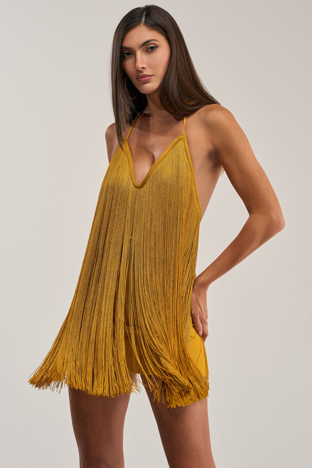 A woman models the DRINKS ON ME FRINGE MINI DRESS IN GOLD, a mustard yellow mini with thin straps and a deep V-neckline, against a plain light background.