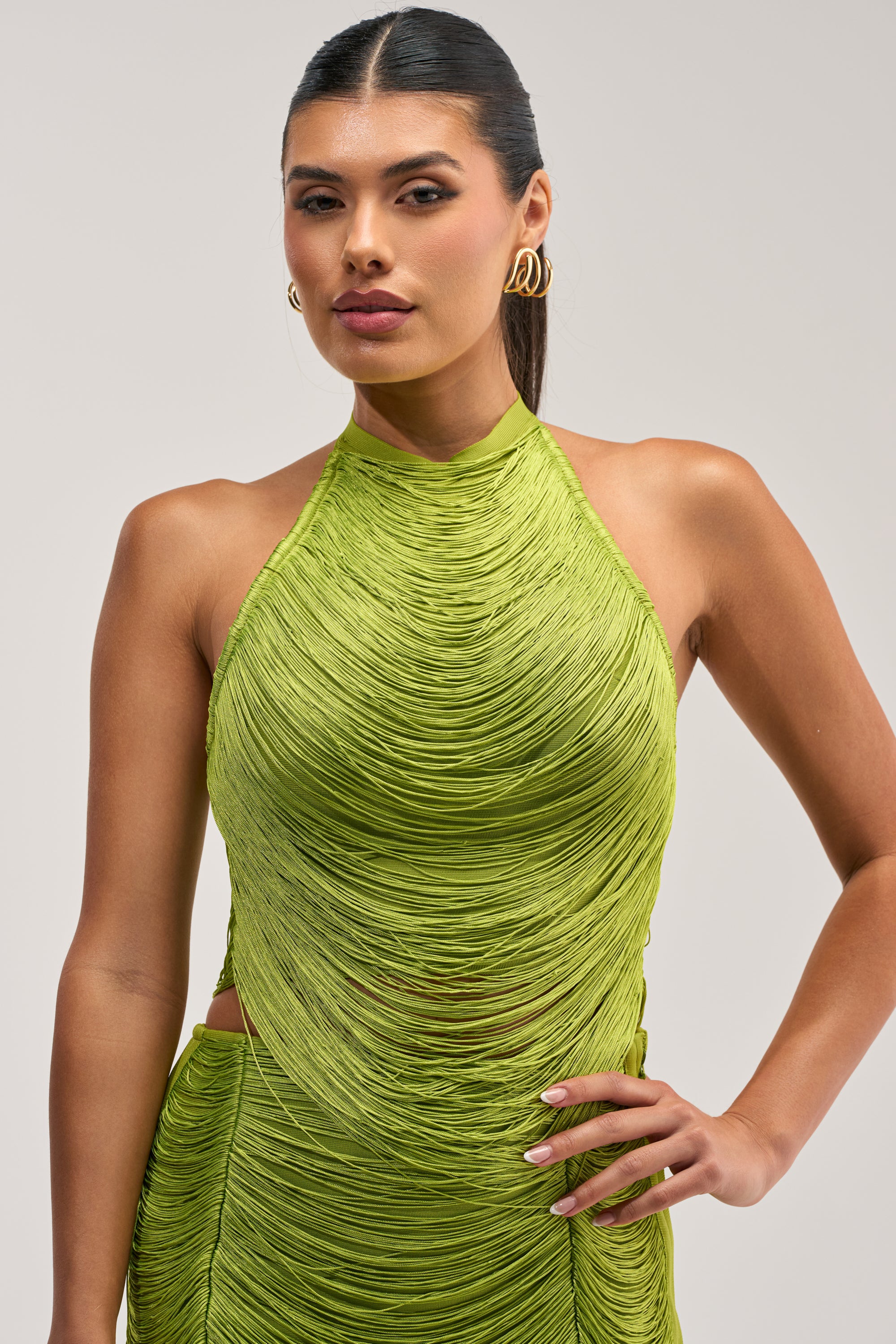 A woman with straight, dark hair pulled back poses against a plain background wearing gold hoop earrings and the BRIGHT AND TIGHT FRINGE TOP—a chartreuse, halter-style bandage top with textured fringe, perfect for birthday styling or updating your look when paired with the matching fringe mini skirt.