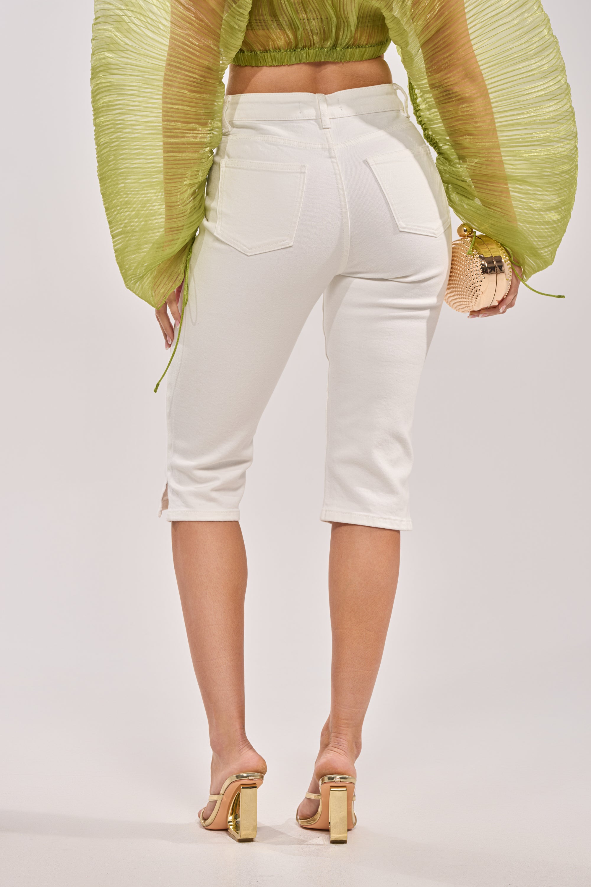 A person wearing CICI DENIM CAPRI in white, a green textured long-sleeve top, gold heeled sandals, and holding a small gold purse is shown from the back against a light background. 3