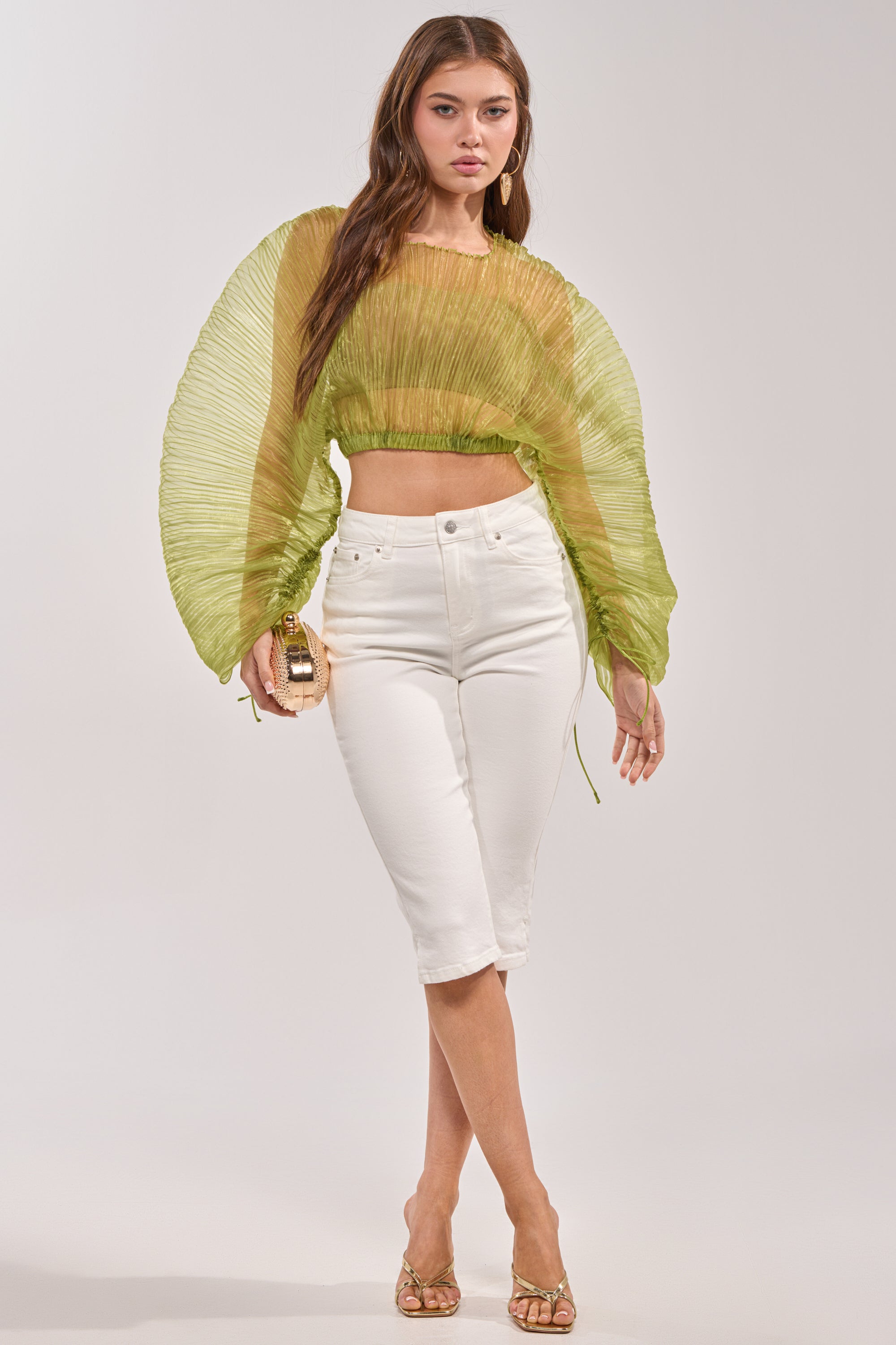 A woman poses against a plain background in a sheer pleated light green crop top, strappy heels, and holds a small tan handbag while wearing the CICI DENIM CAPRI. 4