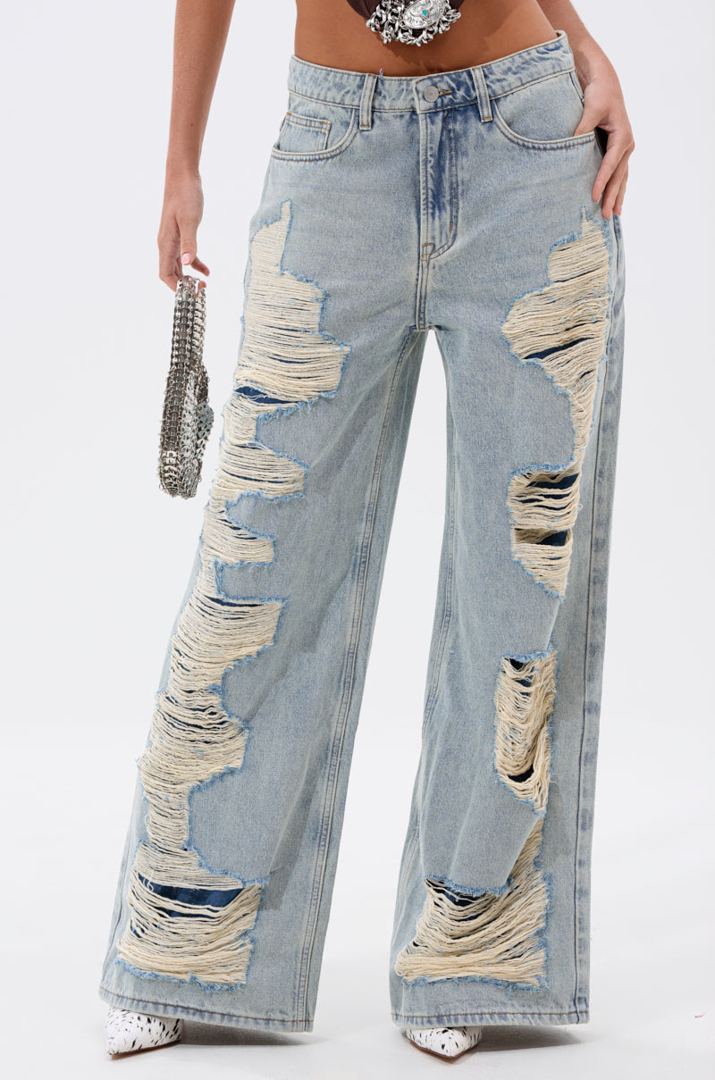 Wearing the CHILL WITH ME DISTRESSED DENIM, featuring light-wash baggy jeans with bold shredded rips revealing beige threads, styled with a silver beaded handbag and white pointed shoes. 0