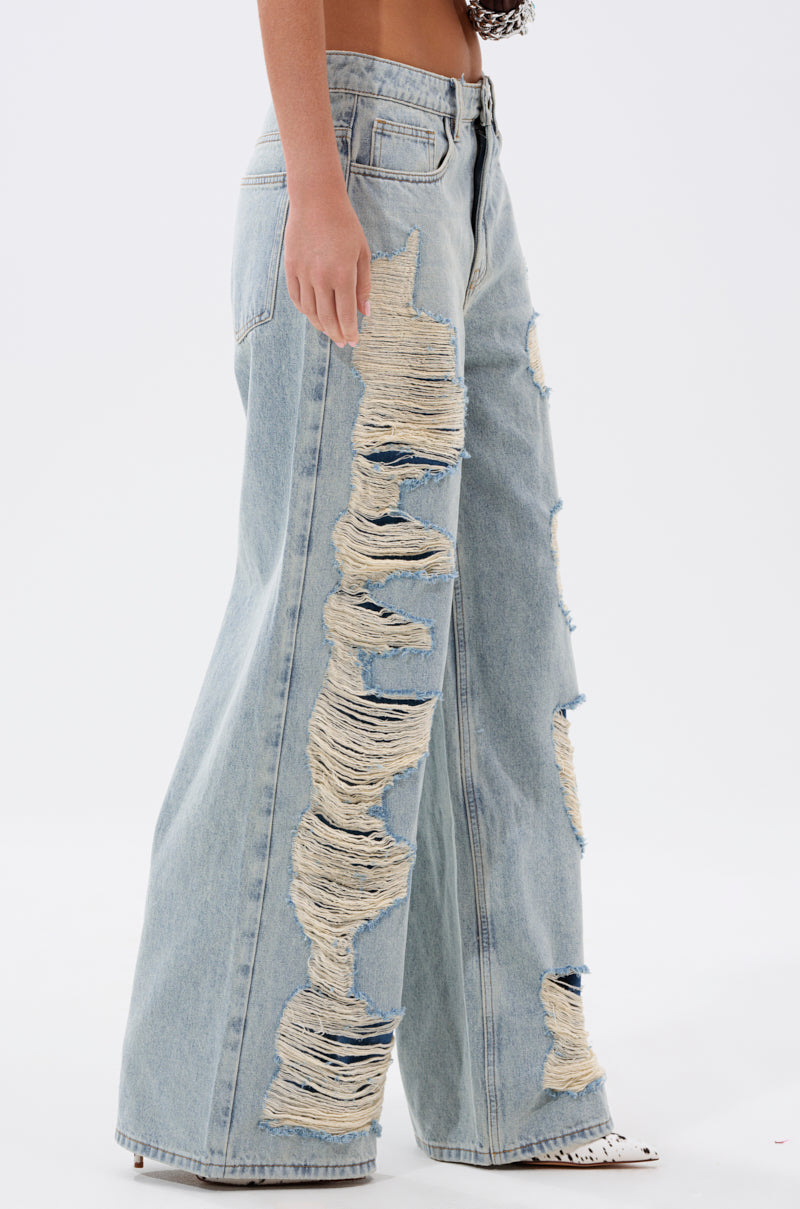 Someone wears the CHILL WITH ME DISTRESSED DENIM, featuring light-wash, wide-leg jeans with large frayed patches along the side seams and front, paired with a cropped top against a plain white background. 2