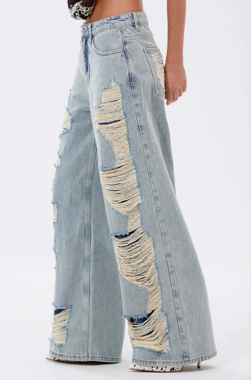 A person models the CHILL WITH ME DISTRESSED DENIM: high-waisted, light-wash baggy jeans with large shredded rips and frayed white threads, photographed from the side against a plain background. 3