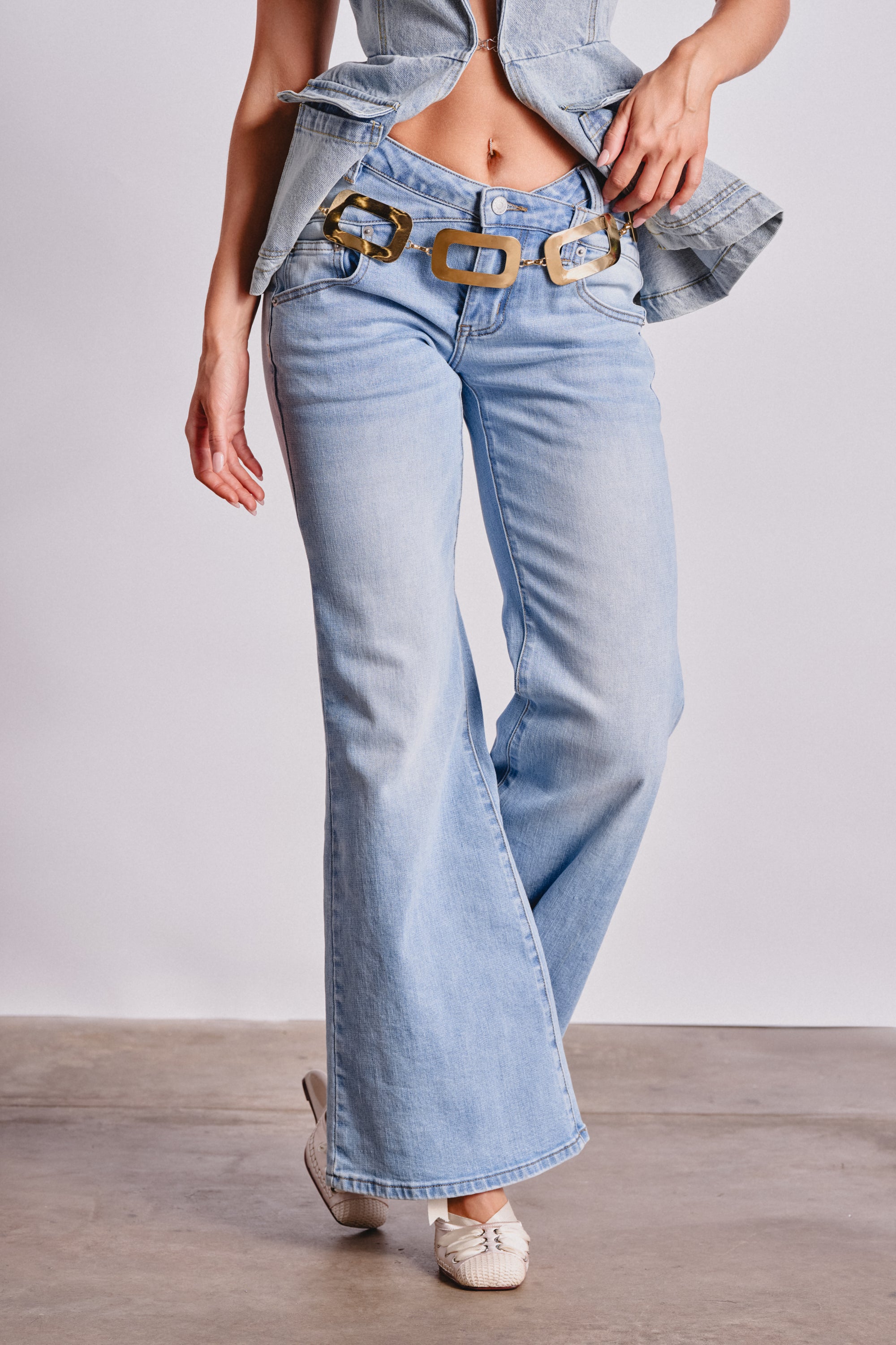 A person wears WON'T LOSE MYSELF WIDE-LEG BLUE DENIM jeans with a denim top, gold chain belt, and white flats, standing on a concrete floor against a plain light background. 0