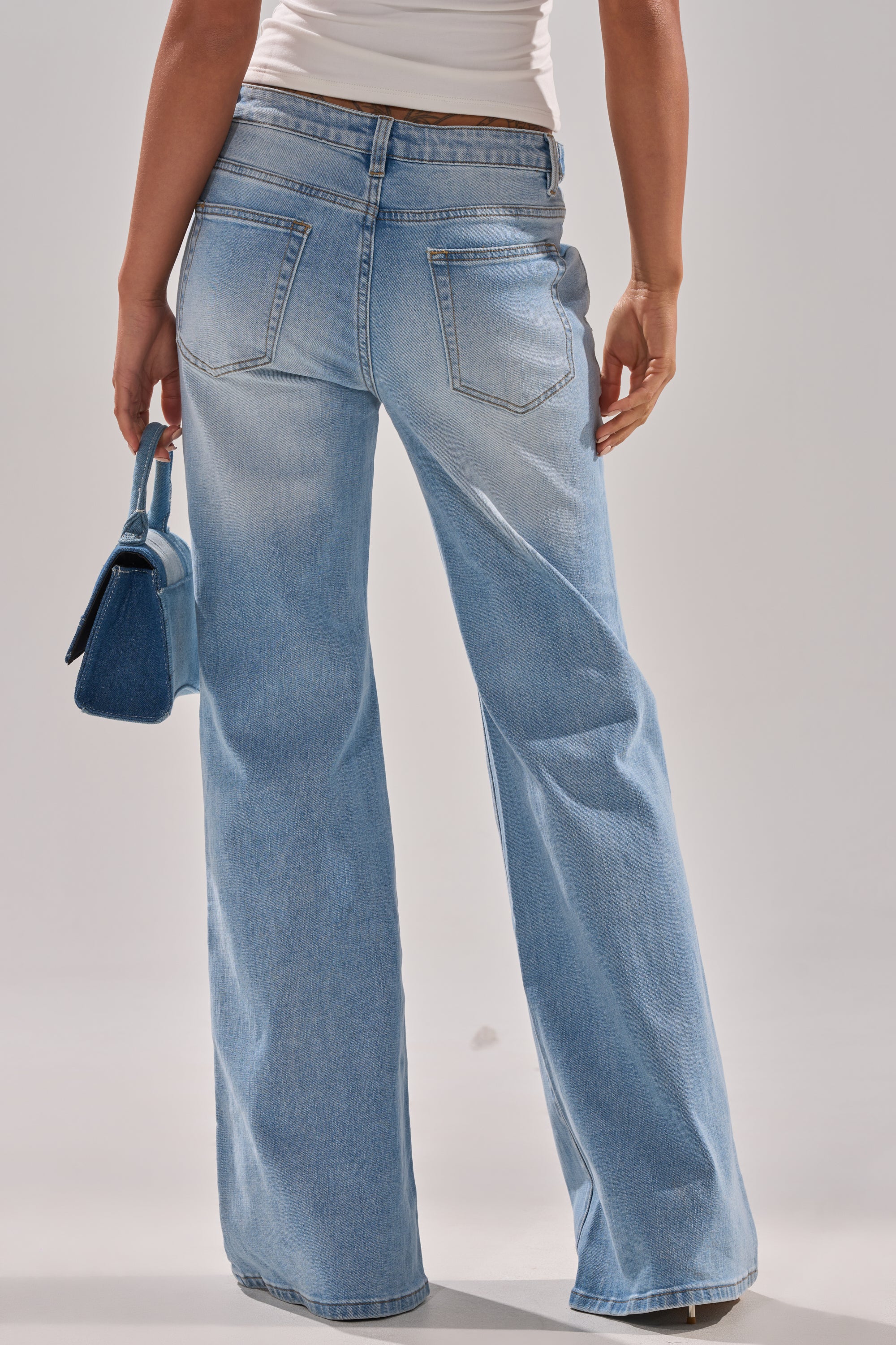 Wearing the WON'T LOSE MYSELF WIDE-LEG BLUE DENIM jeans and a white top, a person stands facing away with a small blue handbag against a plain, light background. 5